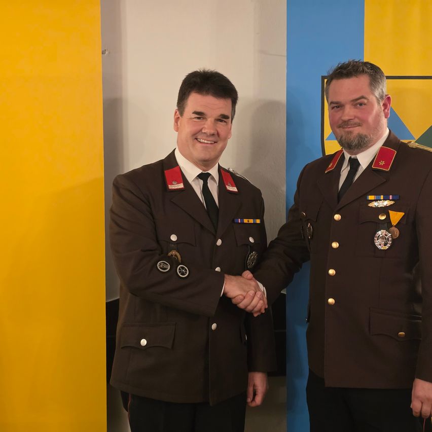 Zwei Männer in Militäruniformen lächeln und schütteln sich die Hände vor einer gelben und blauen Wand.