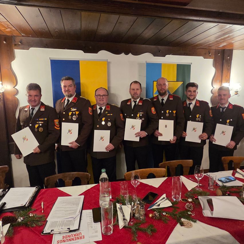 Sieben Männer in Militäruniformen stehen vor einem dekorierten Tisch und halten Urkunden. Dahinter hängen zwei Flaggen, eine blau und gelb und die andere grün und gelb.