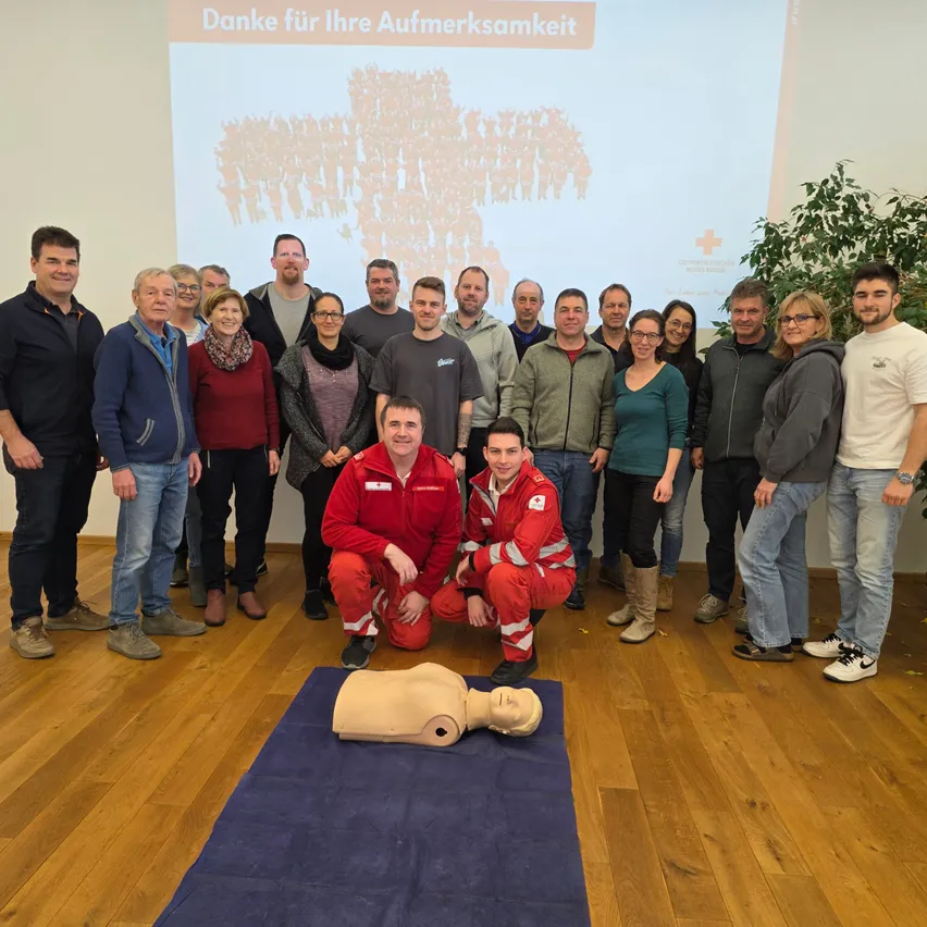 Gruppe von Menschen in einem Raum, zwei knien vor einem CPR-Dummy und lächeln, mit einer Projektionsleinwand im Hintergrund.