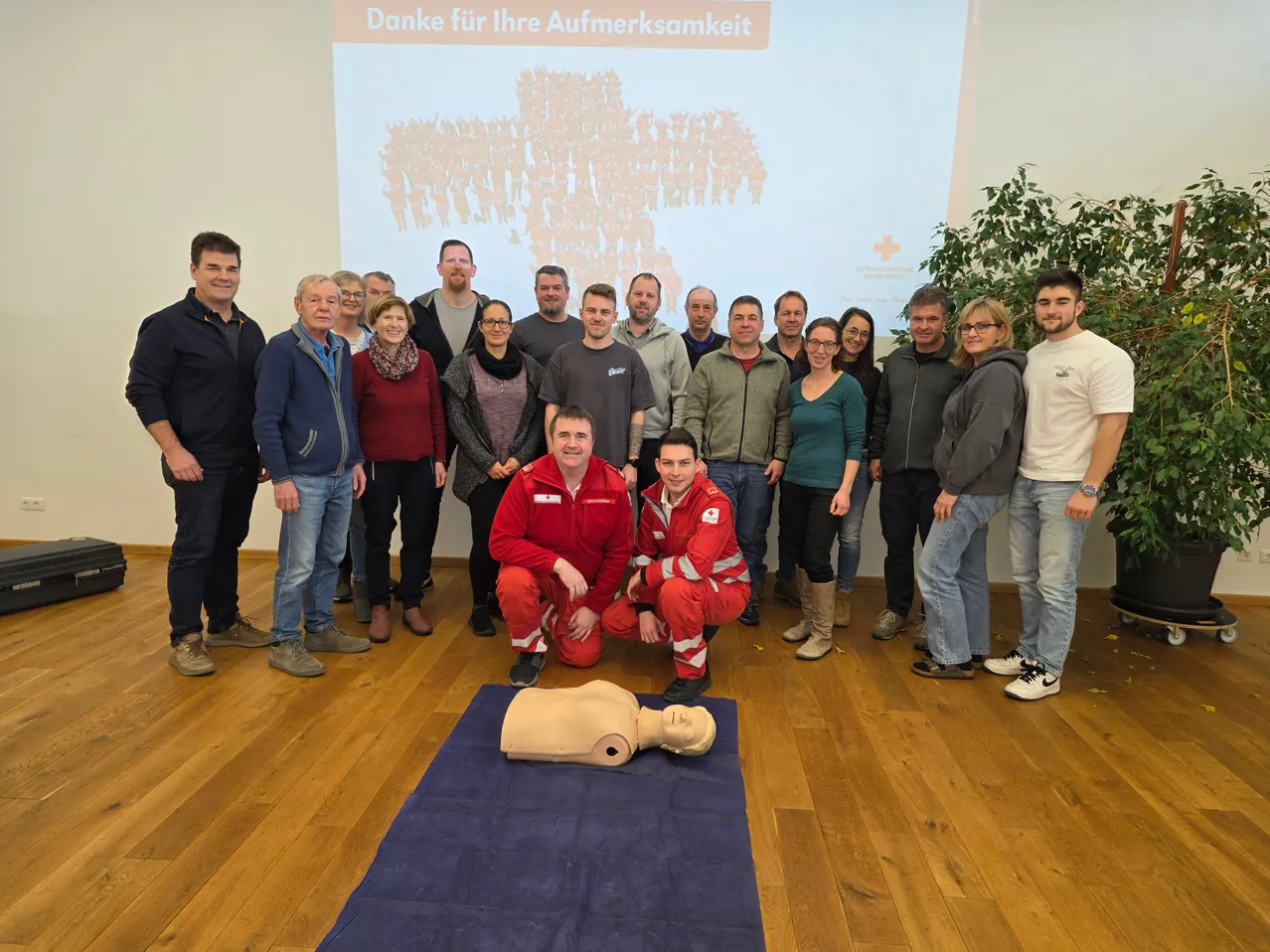 Gruppe von Menschen in einem Raum, zwei knien vor einem CPR-Dummy und lächeln, mit einer Projektionsleinwand im Hintergrund.