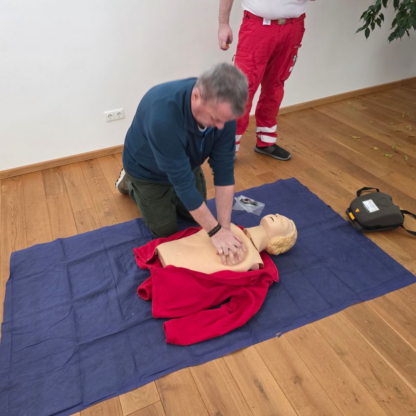 Ein Mann demonstriert CPR an einem Mannequin, das auf einem blauen Teppich in einem Raum mit Holzboden liegt. Eine andere Person steht hinter ihm.