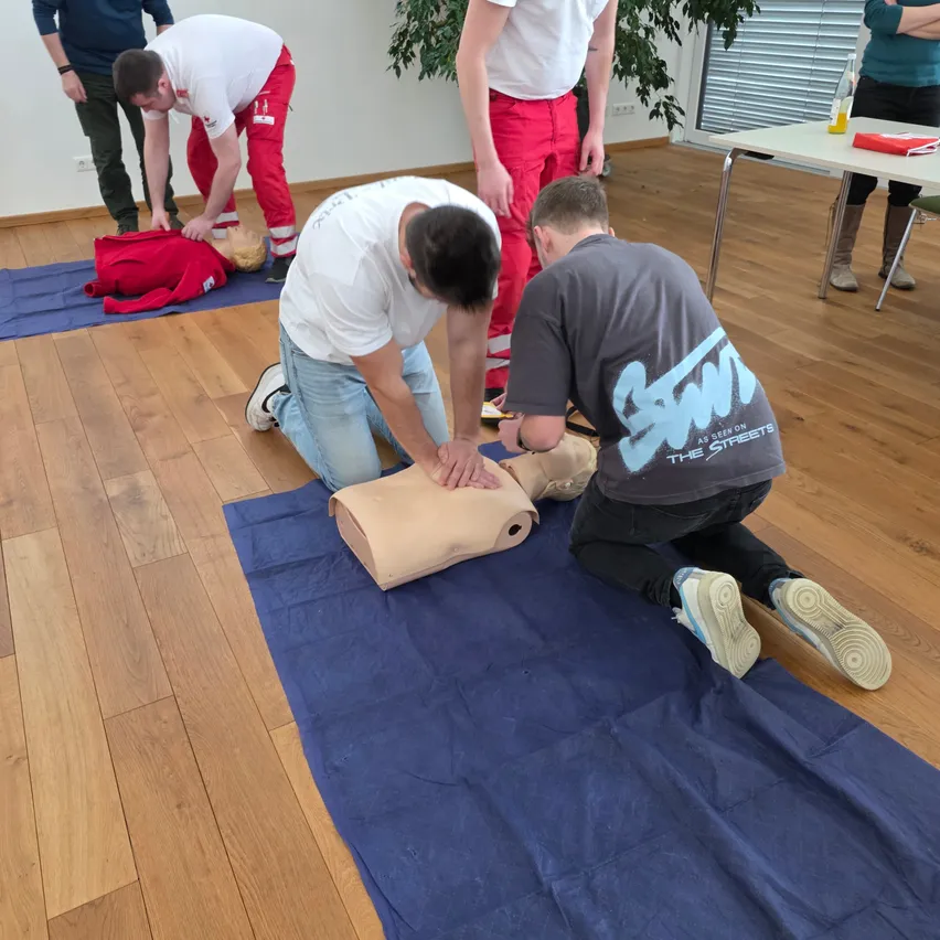 Drei Männer üben CPR an einem Dummies auf einem blauen Teppich, während ein vierter Mann steht und beobachtet. Dahinter stellt ein weiterer eine Person am Dummies auf einem anderen Teppich ein. Es gibt einen Tisch mit Papieren und einer Flasche.