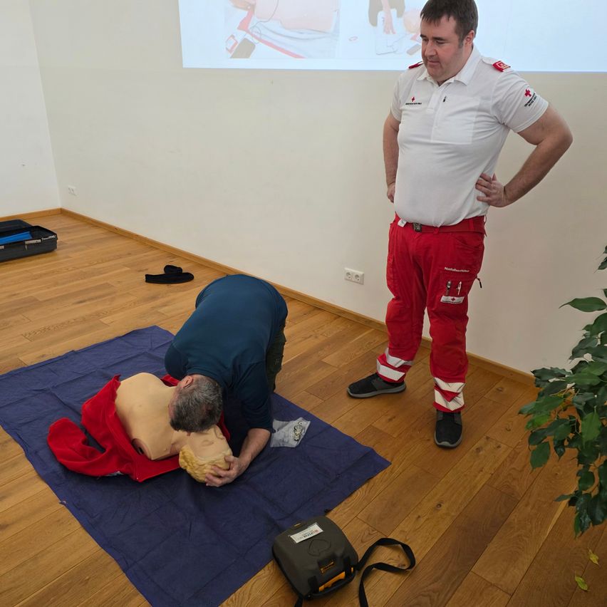Ein Mann übt CPR auf einem Dummies auf einer blauen Matte, während ein anderer Mann in roten Hosen zuschaut. Eine Projektionsleinwand zeigt ein Bild im Hintergrund.