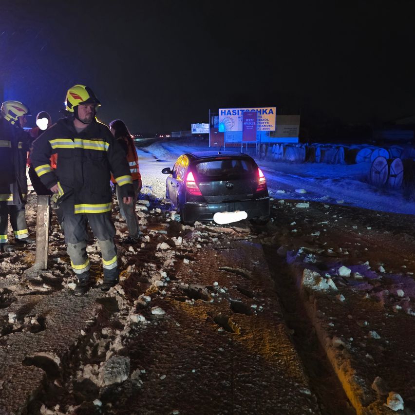 Feuerwehrleute stehen auf einer mit Steinen bedeckten Straße. Ein beschädigtes Auto ist hinter ihnen geparkt. Die Szene wird von Scheinwerfern und Straßenlaternen beleuchtet.