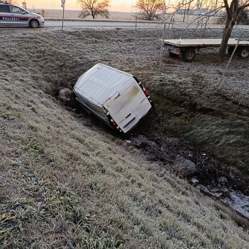 Ein Van steckt in einem Graben in der Nähe einer Straße mit einem Polizeiauto und Anhänger in der Nähe, umgeben von Bäumen und Gras mit einem Sonnenuntergang im Hintergrund.