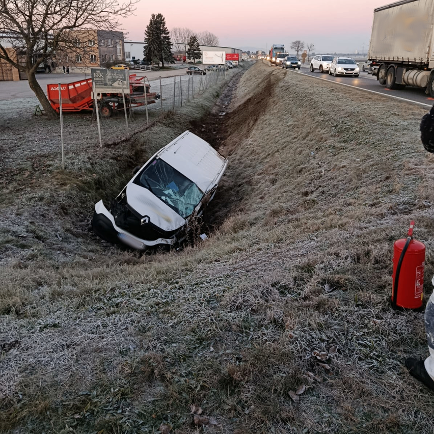 Ein demoliertes Fahrzeug steckt im Straßengraben, mit einem Feuerlöscher in der Nähe. Eine Autobahn verläuft parallel zu einem Gebäude und Bäumen im Hintergrund.
