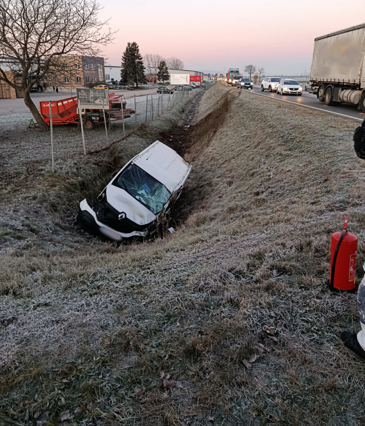 Ein demoliertes Fahrzeug steckt im Straßengraben, mit einem Feuerlöscher in der Nähe. Eine Autobahn verläuft parallel zu einem Gebäude und Bäumen im Hintergrund.