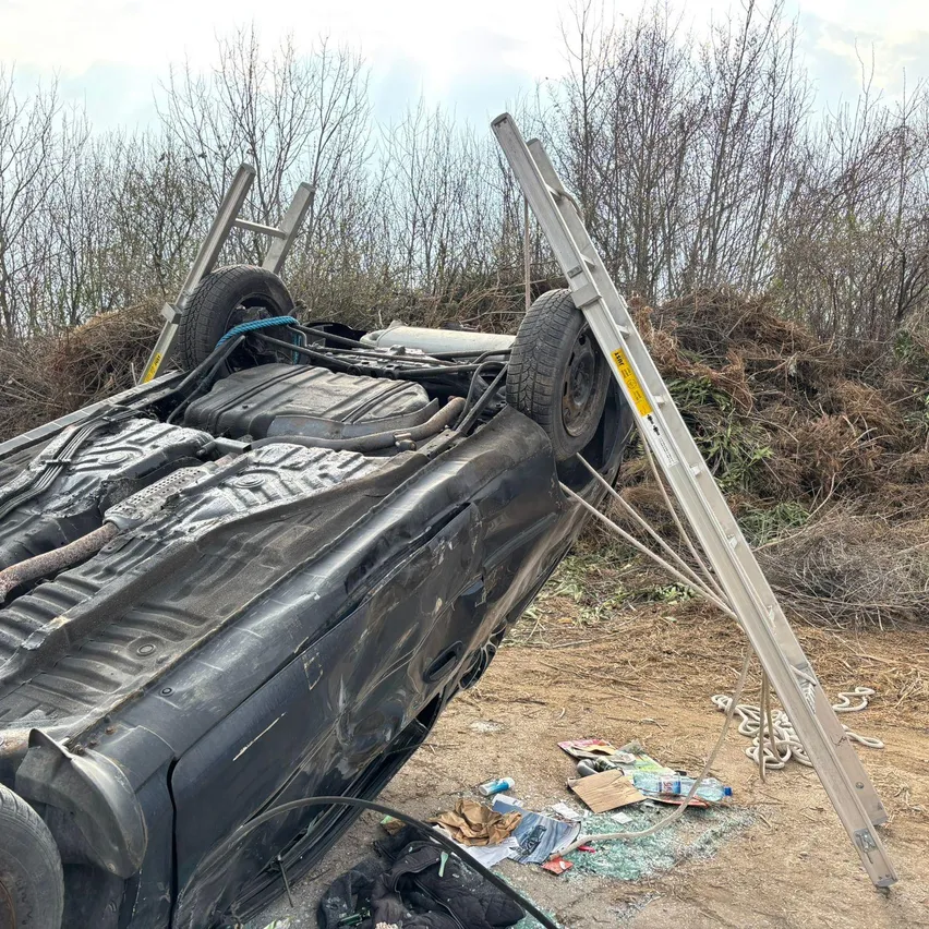 Ein umgekipptes Auto mit der Front nach oben, gesichert durch Leitern und Seile. Die Szene umfasst Schutt und einen schlammigen Boden.