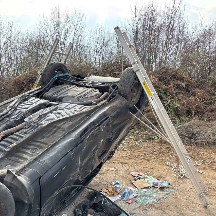 Ein umgekipptes Auto mit der Front nach oben, gesichert durch Leitern und Seile. Die Szene umfasst Schutt und einen schlammigen Boden.