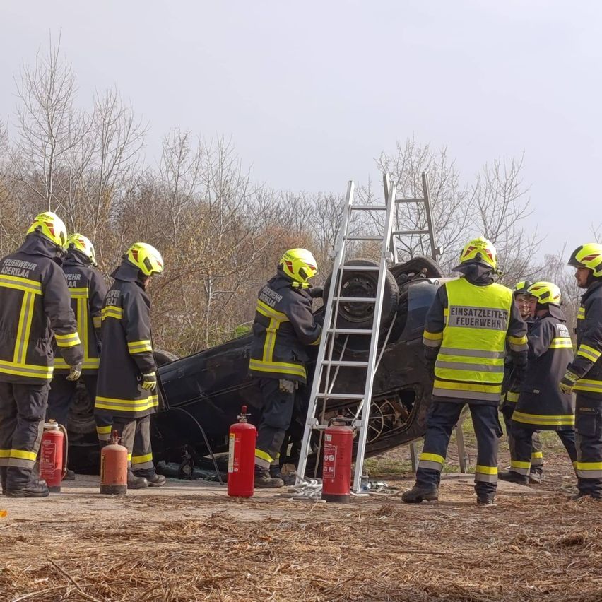 Eine Gruppe von Feuerwehrleuten in gelben Helmen arbeitet an einem umgekippten Auto mit einer Leiter. Sie sind von Feuerlöschern umgeben.