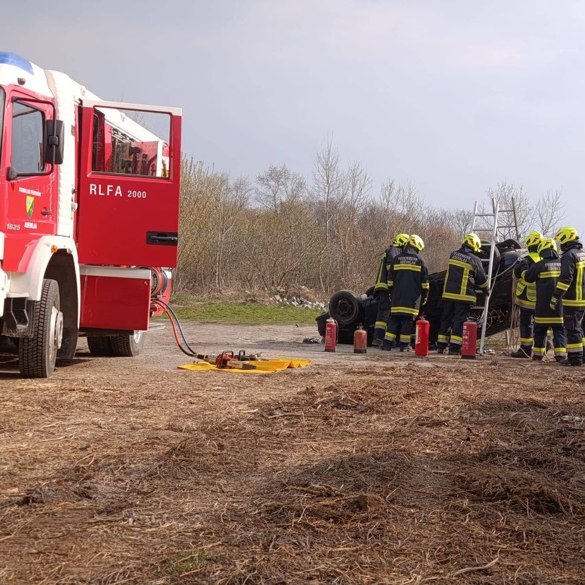 Ein roter Feuerwehrwagen mit der Aufschrift RLFA 2000 ist im Freien geparkt. Feuerwehrleute in gelben Helmen stehen in der Nähe, mit Feuerlöschern auf dem Boden platziert.