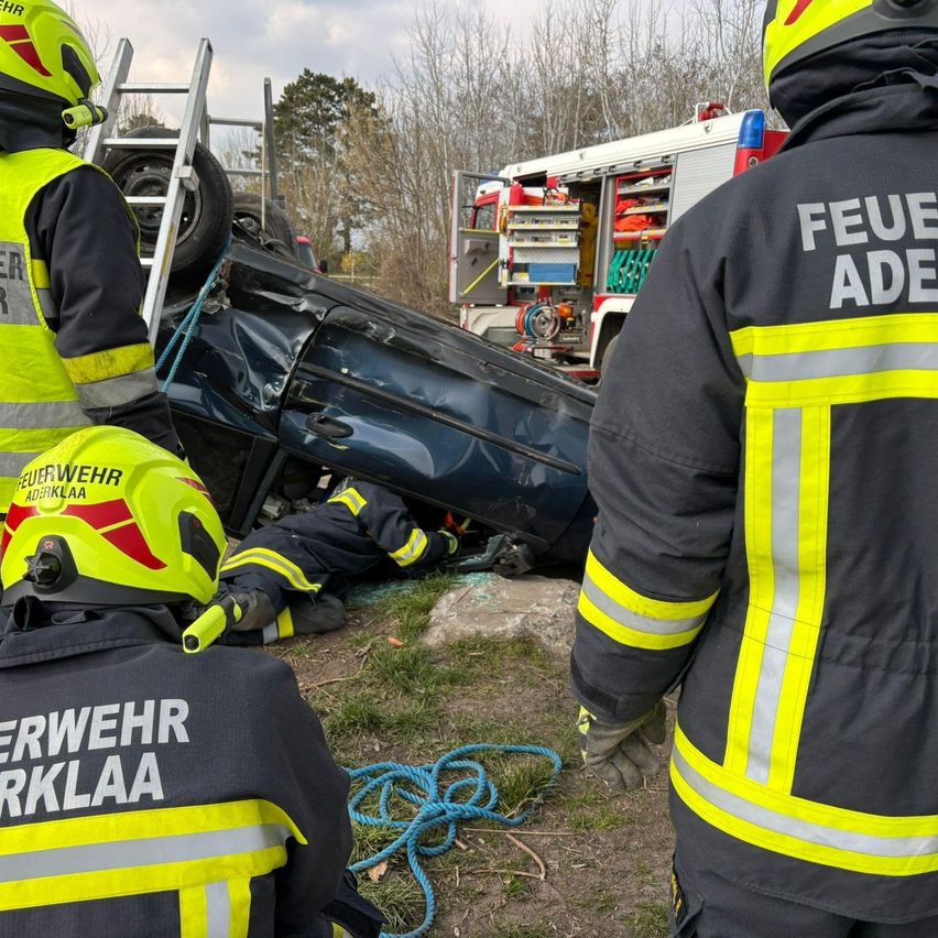 Rettungskräfte in gelben und schwarzen Uniformen kümmern sich um ein zerstörtes Auto am Straßenrand. Ein Feuerwehrmann steht neben dem Fahrzeug.