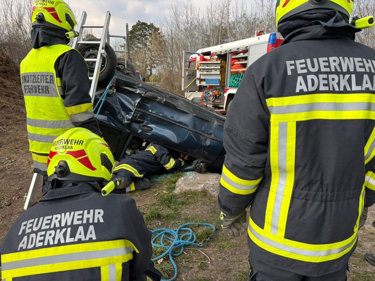 Rettungskräfte in gelben und schwarzen Uniformen kümmern sich um ein zerstörtes Auto am Straßenrand. Ein Feuerwehrmann steht neben dem Fahrzeug.