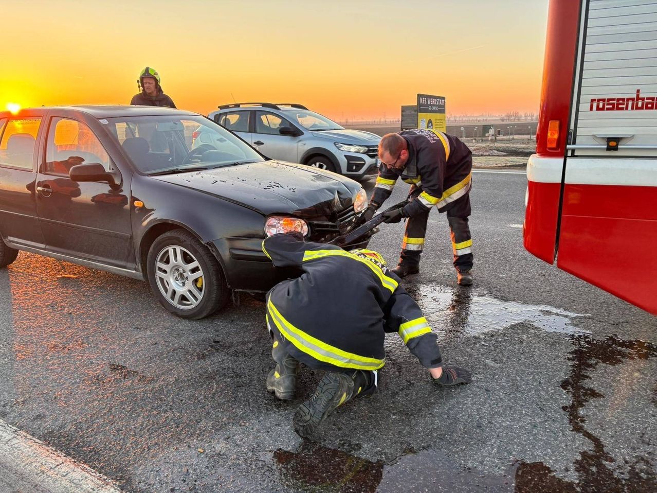 Zwei Feuerwehrleute arbeiten an einem beschädigten schwarzen Auto am Straßenrand. Eine weitere Person steht in der Nähe, und ein Feuerwehrwagen ist dahinter geparkt.