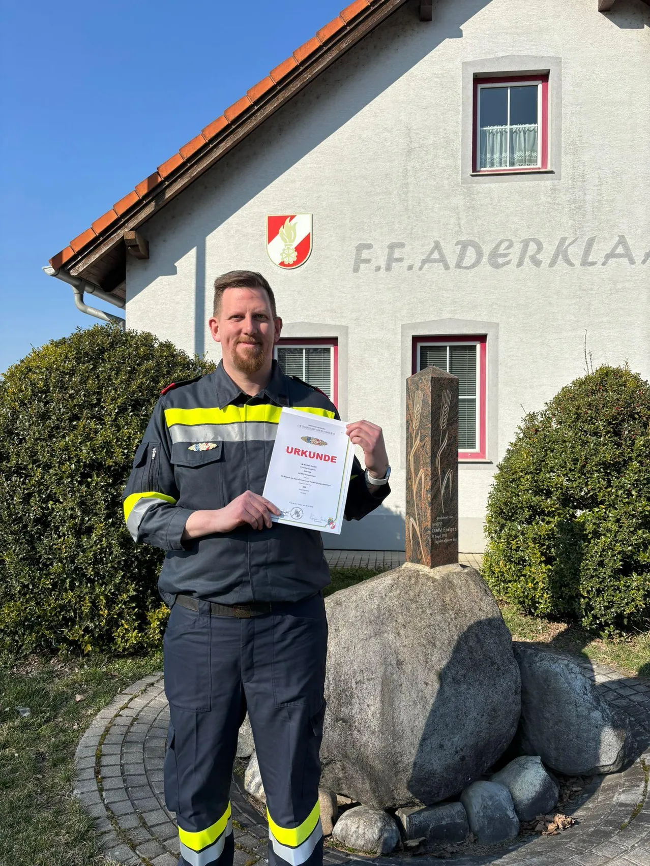 Ein Mann steht vor einem weißen Gebäude mit einem roten Logo und hält ein Zertifikat mit der Aufschrift 'URKUNDE'. Er steht vor einem großen Felsen mit einem kleinen Steinobelisk.