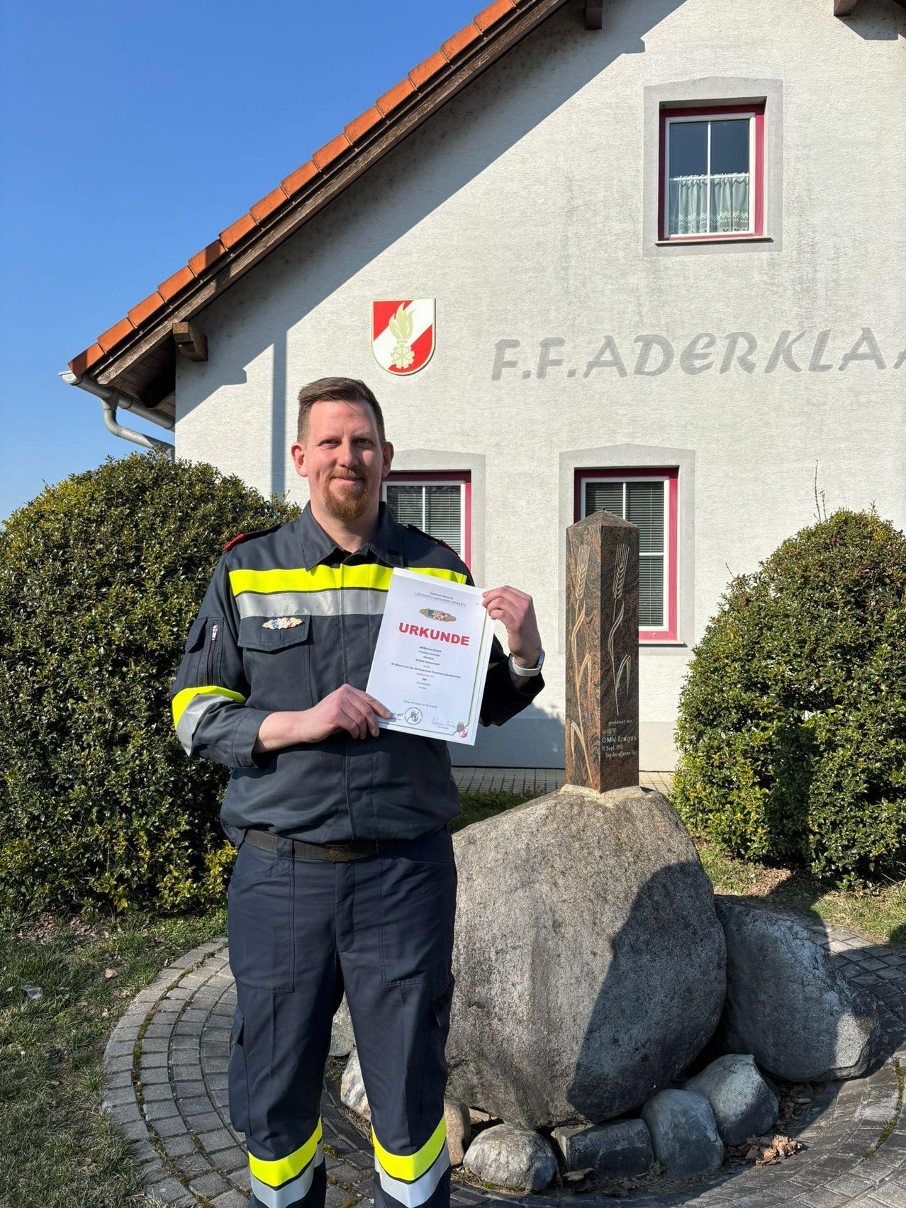 Ein Mann steht vor einem weißen Gebäude mit einem roten Logo und hält ein Zertifikat mit der Aufschrift 'URKUNDE'. Er steht vor einem großen Felsen mit einem kleinen Steinobelisk.