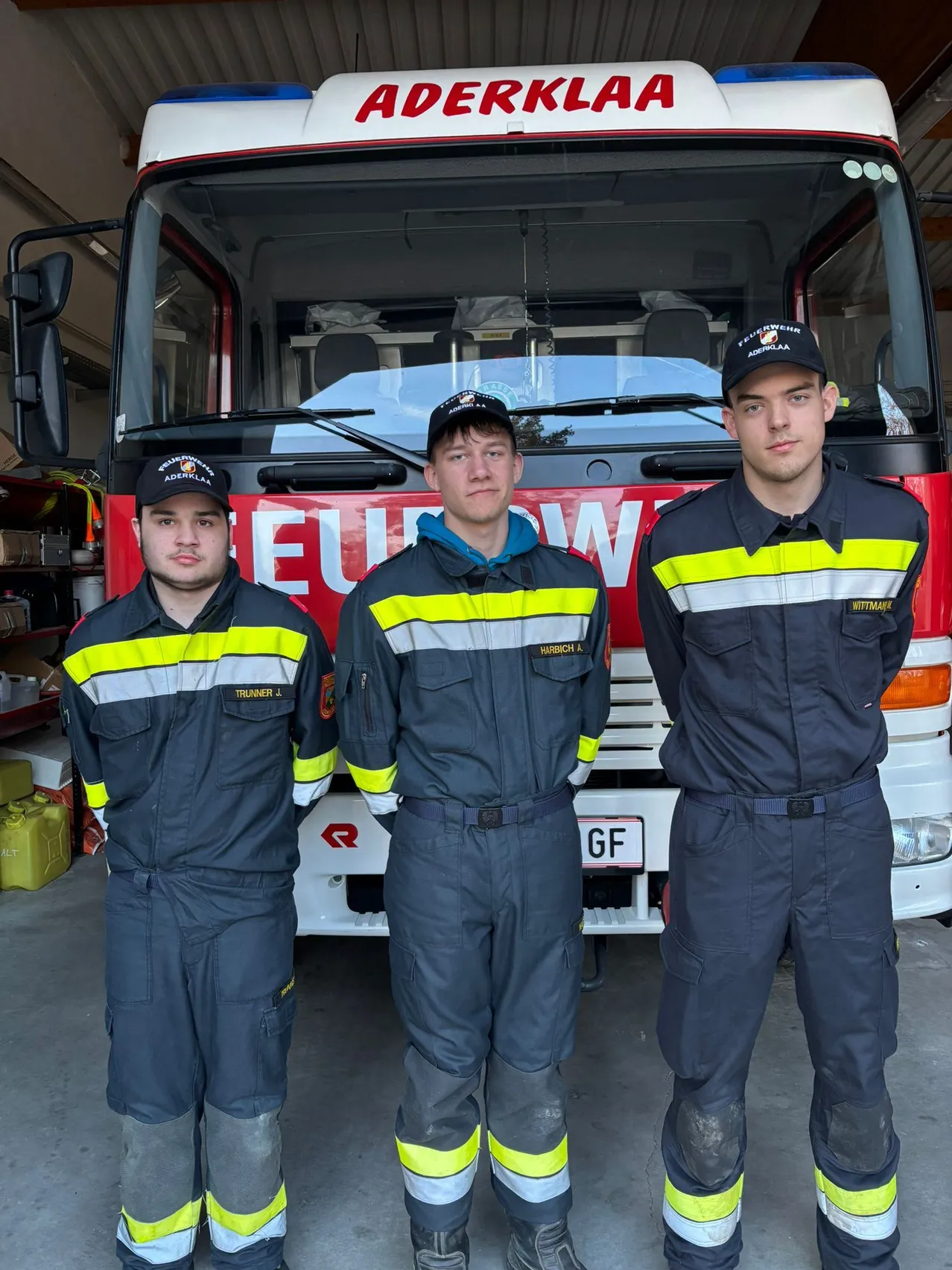 Drei Feuerwehrleute in Uniform stehen vor einem Feuerwehrauto, wahrscheinlich für ein Foto. Das Feuerwehrauto ist rot mit einem weißen Streifen und dem Wort 'EUW' auf der Vorderseite.