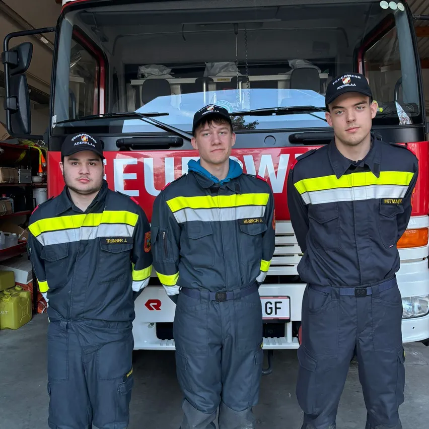 Drei Feuerwehrleute in Uniform stehen vor einem Feuerwehrauto, wahrscheinlich für ein Foto. Das Feuerwehrauto ist rot mit einem weißen Streifen und dem Wort 'EUW' auf der Vorderseite.