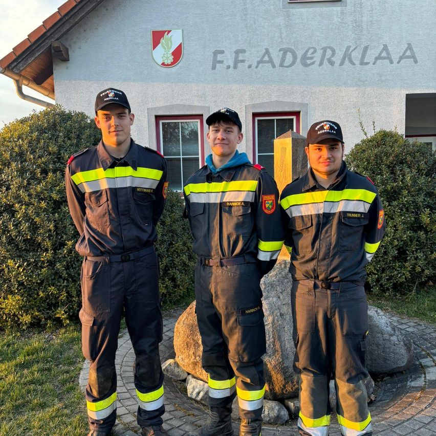 Drei Feuerwehrleute stehen vor einem Gebäude mit 'F.F.Aderklaa' an der Wand. Der Mann links trägt einen 'Wittmar'-Abzeichen, der mittlere Mann ein 'Harrich A'-Abzeichen und der Mann rechts ein 'Trummer J'-Abzeichen.