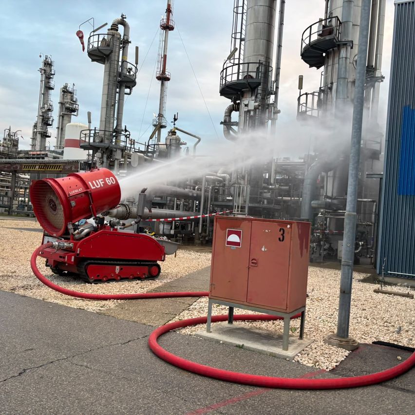 Eine rote Feuerlöscheinheit sprüht Wasser in einem Industriegebiet mit großen Rohren und Tanks im Hintergrund.