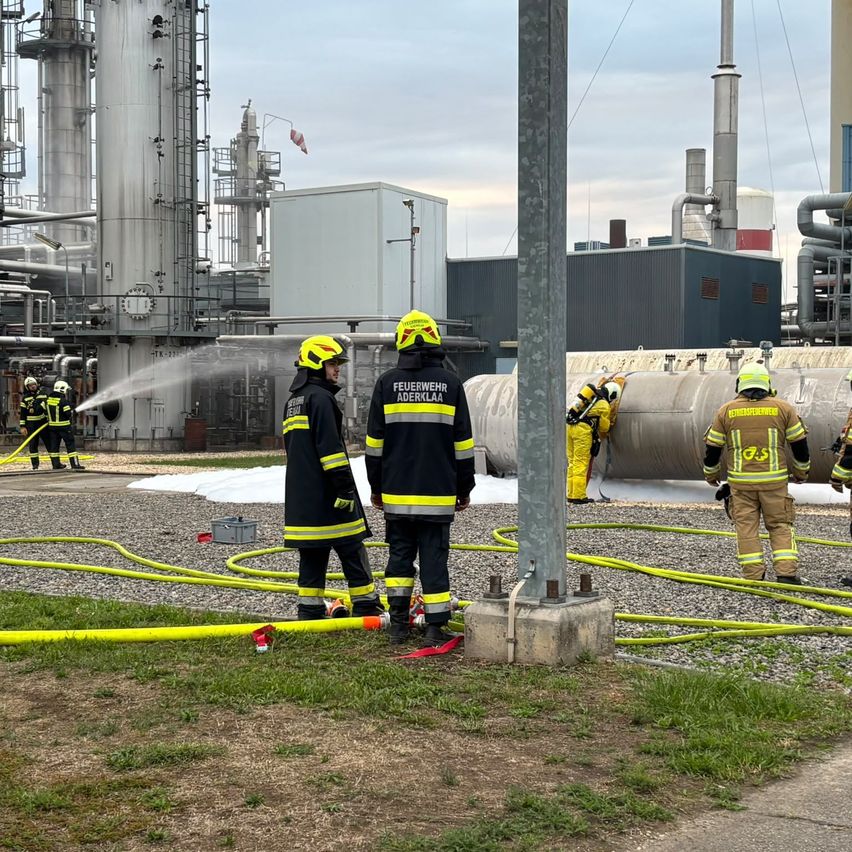 Feuerwehrleute in Schutzkleidung stehen vor einer großen Industrieanlage, einer spritzt Wasser aus einem Schlauch.