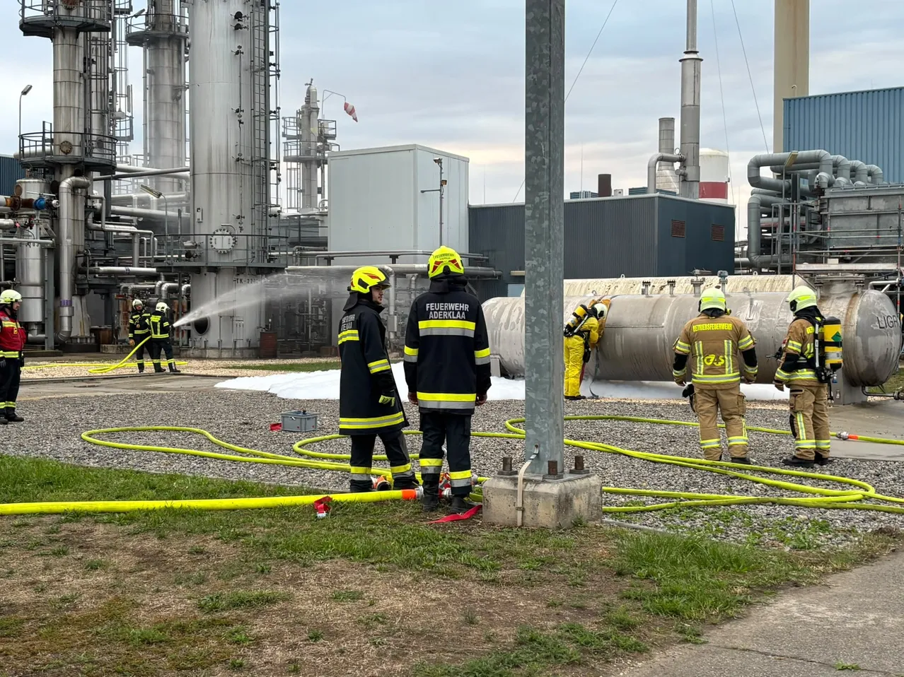 Feuerwehrleute in Schutzkleidung stehen vor einer großen Industrieanlage, einer spritzt Wasser aus einem Schlauch.