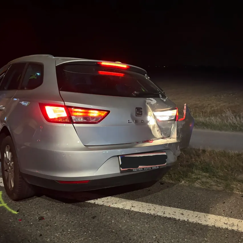 Ein silbernes Auto ist nachts am Straßenrand geparkt. Die hintere Tür ist offen und die Rücklichter sind eingeschaltet.