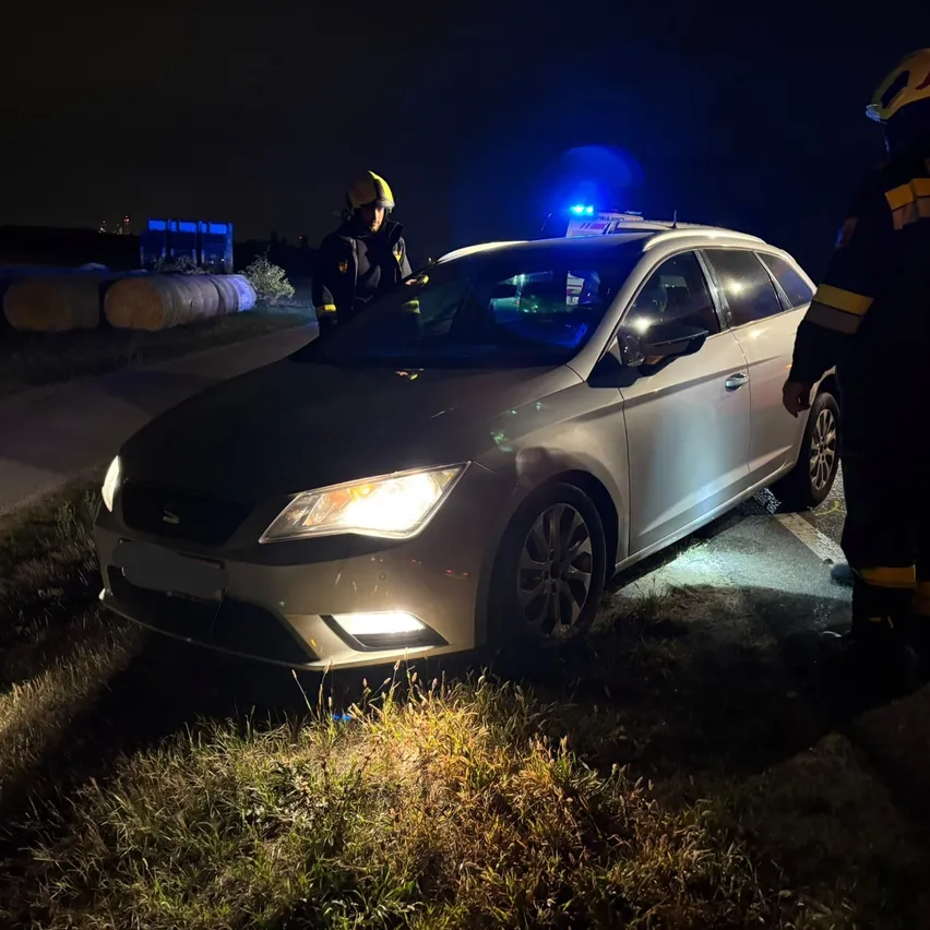 Zwei Feuerwehrleute kümmern sich bei Nacht um einen silbernen Wagen am Straßenrand. Die Scheinwerfer des Autos sind eingeschaltet, und einer der Feuerwehrleute steht neben der offenen Fahrertür.