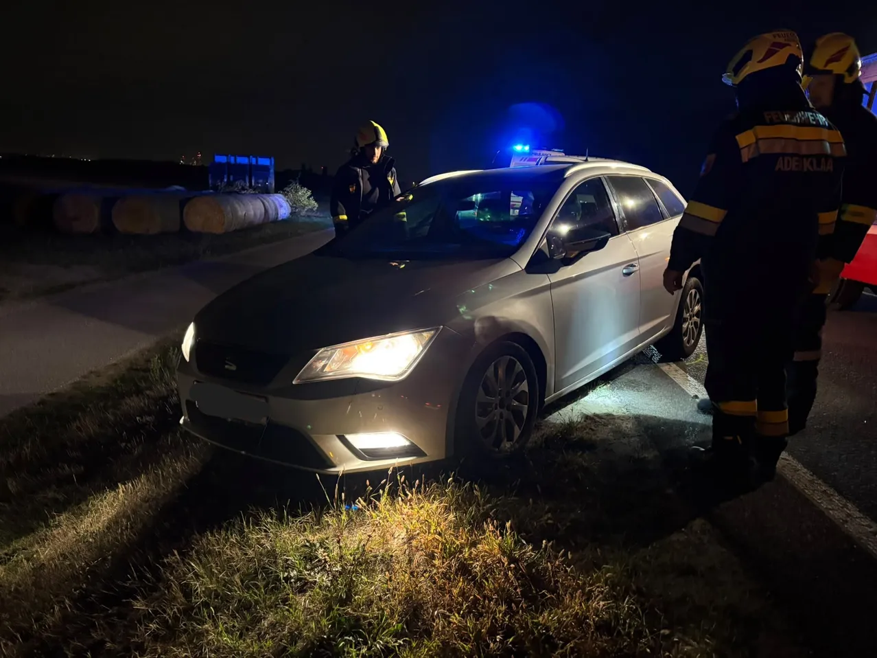 Zwei Feuerwehrleute kümmern sich bei Nacht um einen silbernen Wagen am Straßenrand. Die Scheinwerfer des Autos sind eingeschaltet, und einer der Feuerwehrleute steht neben der offenen Fahrertür.