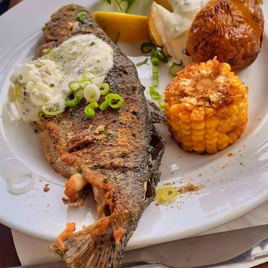 Ein Teller mit gebratenem Fisch, cremiger Sauce, grünen Zwiebeln, einer gebackenen Kartoffel mit Sauerrahm und einem Maiskolben.