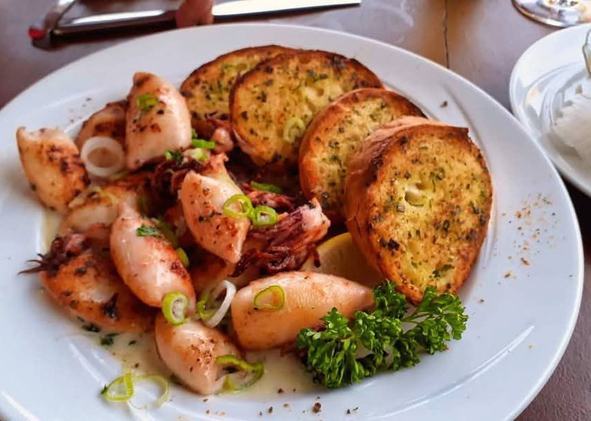 Ein weißer Teller mit gegrilltem Oktopus, Knoblauchbrot und Petersiliengarnitur. Der Oktopus ist geschnitten und mit Kräutern gewürzt.