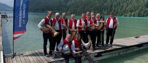 Eine Gruppe von Musikern in roten Westen steht auf einer Pier am See mit Bergen im Hintergrund. Sie halten Blechblasinstrumente.