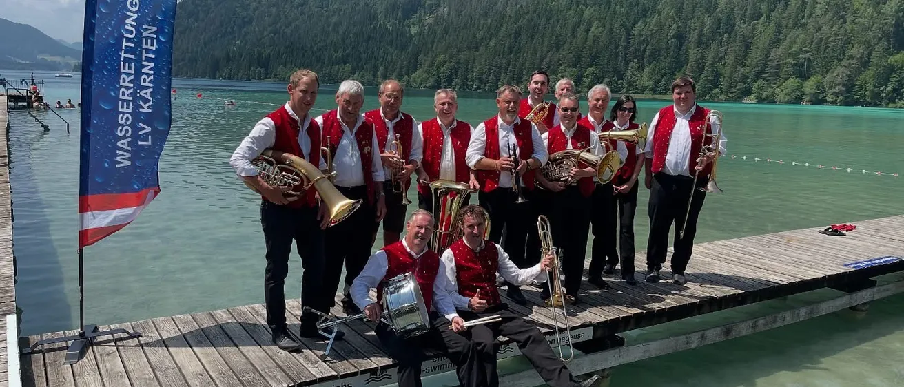 Eine Gruppe von Musikern in roten Westen steht auf einer Pier am See mit Bergen im Hintergrund. Sie halten Blechblasinstrumente.