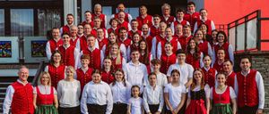 Eine große Gruppe von Menschen in traditioneller deutscher Kleidung steht auf Stufen und lächelt für ein Foto. Sie besteht aus Männern und Frauen, einige tragen rote Westen mit weißen Punkten.