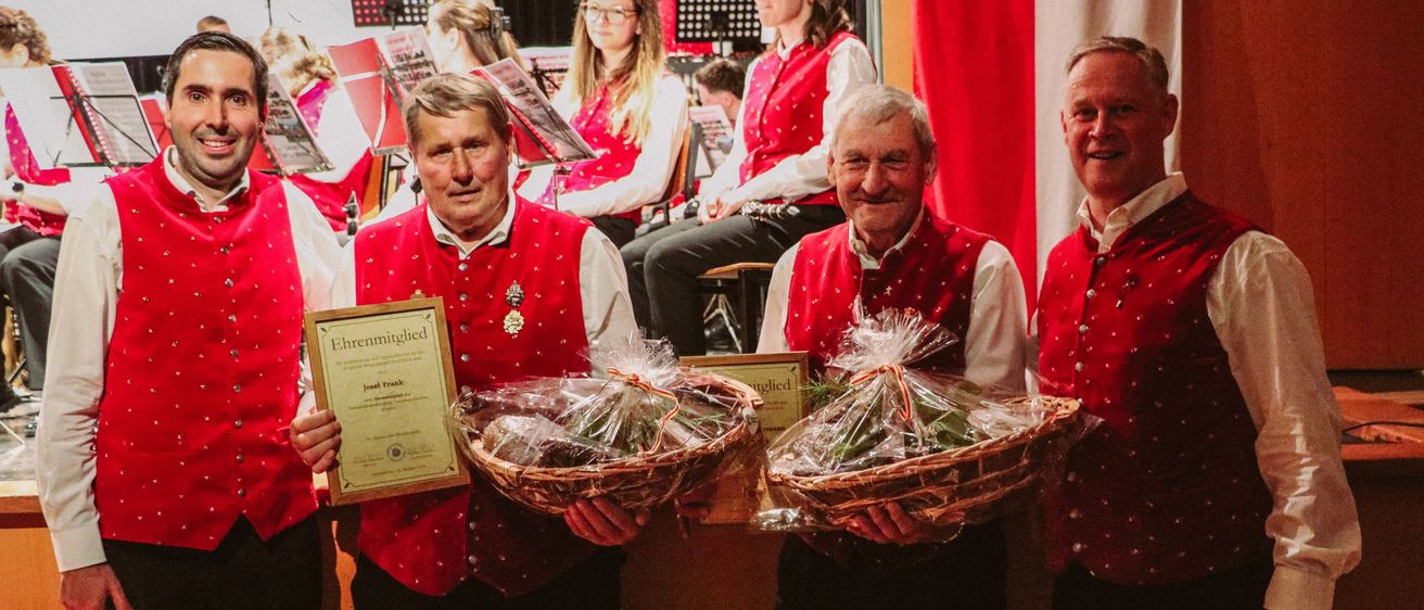 Fünf Männer in roten Westen und schwarzen Hosen halten einen Korb mit einem Zertifikat. Sie lächeln und posieren für ein Foto. Hinter ihnen ist ein unscharfer Hintergrund einer Bühne und eines roten Vorhangs.