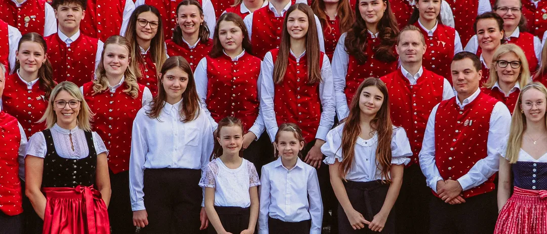 Ein Chor in roten Westen und schwarzen Hosen posiert für ein Gruppenfoto auf einer Treppe. Der Chor besteht aus Erwachsenen und Kindern, und alle lächeln.