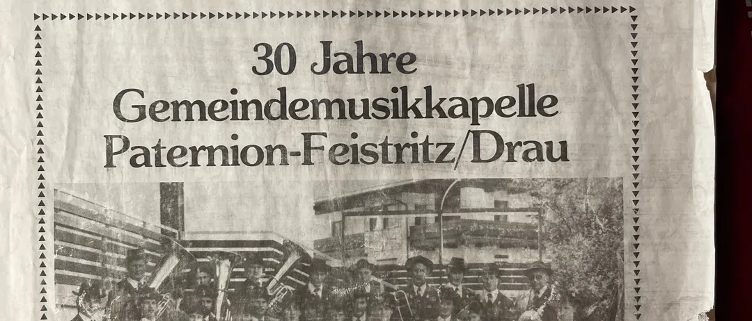 Ein Schwarz-Weiß-Foto einer Blaskapelle mit Mitgliedern in Uniformen und Hüten steht vor einem Gebäude. Es ist eine Seite aus einer Gemeindezeitung für Paterion-Feistritz/Drau, August 1983.
