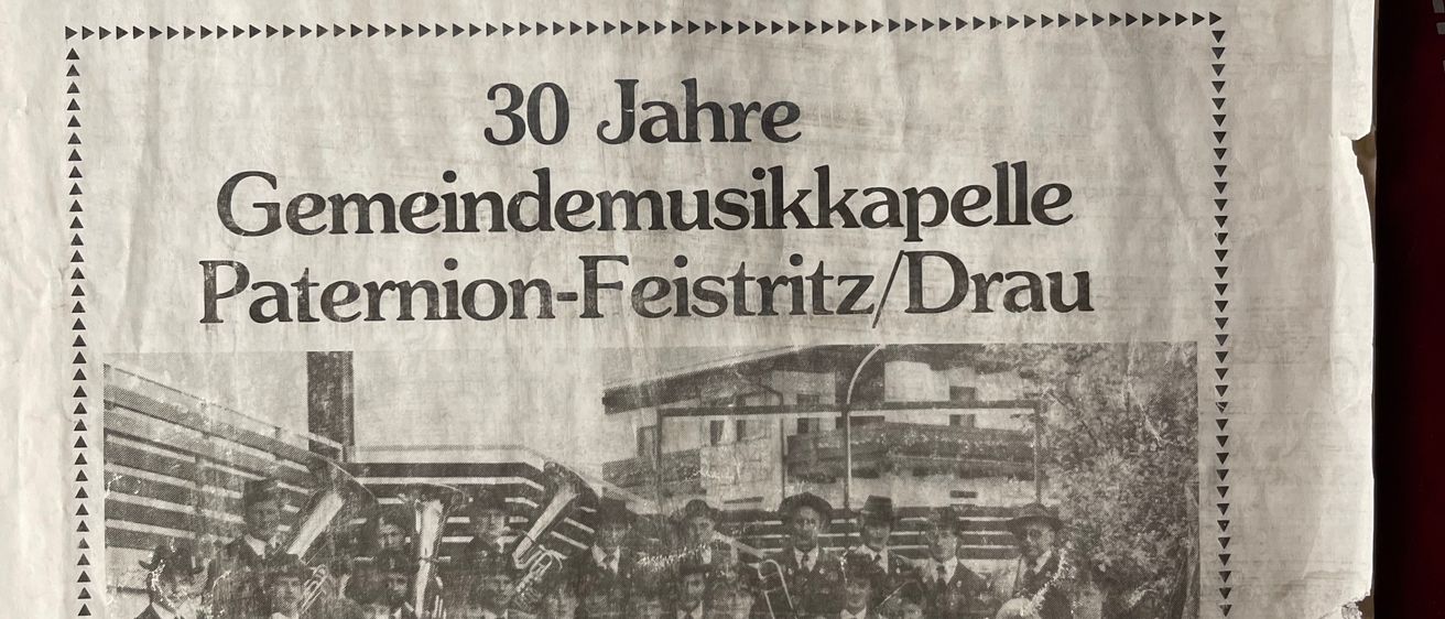 Ein Schwarz-Weiß-Foto einer Blaskapelle mit Mitgliedern in Uniformen und Hüten steht vor einem Gebäude. Es ist eine Seite aus einer Gemeindezeitung für Paterion-Feistritz/Drau, August 1983.