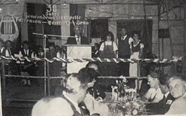 Ein Schwarz-Weiß-Bild zeigt einen Mann, der an einem Podium spricht, mit drei Frauen hinter ihm. Das Publikum sitzt, einige tragen Krawatten. Ein Banner mit deutschem Text ist sichtbar.