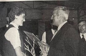 Ein Mann in Anzug überreicht einer Frau in einem weißen Shirt eine Auszeichnung, mit einem Mann in Weste im Hintergrund.