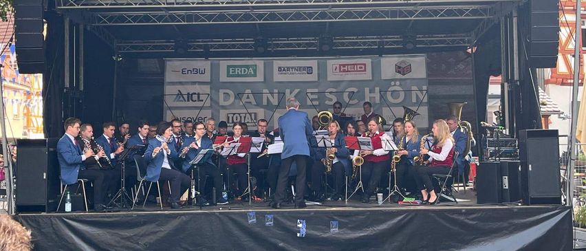 Eine Band spielt auf der Bühne vor einem Gebäude mit rot-weißer Fassade. Die Bühne hat Sponsorenbanner. Die Musiker spielen verschiedene Instrumente, darunter Flöten, Saxophone und Trompeten.