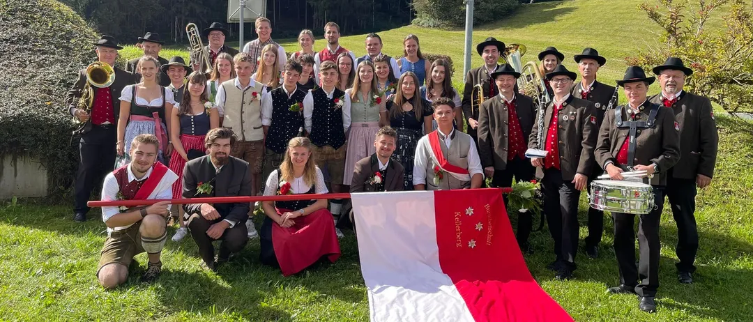 Eine Gruppe von Menschen in traditionellen Kostümen posiert für ein Foto auf einem Rasenfeld mit einer Flagge. Dahinter ein landschaftlicher Blick auf einen Berg und einen Pfosten.