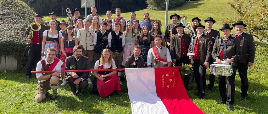 Eine Gruppe von Menschen in traditionellen Kostümen posiert für ein Foto auf einem Rasenfeld mit einer Flagge. Dahinter ein landschaftlicher Blick auf einen Berg und einen Pfosten.