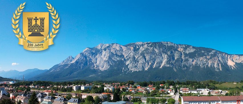 Luftaufnahme einer Stadt mit einem Gebirgszug im Hintergrund. Die Stadt hat viele Gebäude, Bäume und einen klaren blauen Himmel.