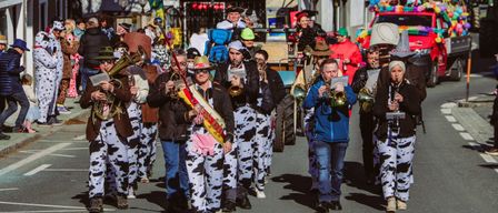Eine Gruppe von Musikern in Kuhkostümen zieht bei einem Umzug mit, mit Zuschauern und Straßendekorationen im Hintergrund.