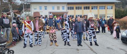 Eine Gruppe von Menschen in Kuhkostümen, darunter zwei Männer mit Trompeten, steht vor einem Gebäude mit einem Banner, auf dem 'Herzlich willkommen' steht.