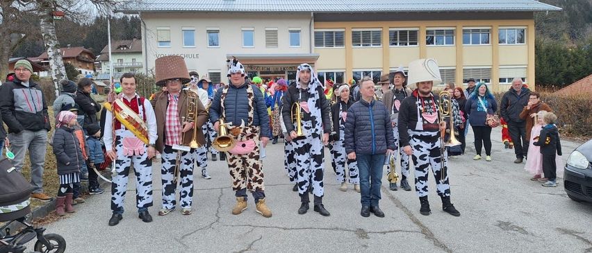 Eine Gruppe von Menschen in Kuhkostümen, darunter zwei Männer mit Trompeten, steht vor einem Gebäude mit einem Banner, auf dem 'Herzlich willkommen' steht.