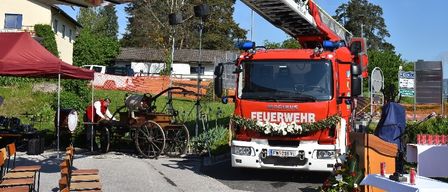 Ein roter Feuerwehrwagen mit ausgefahrener Leiter ist vor einem Haus geparkt, in der Nähe eines Zeltes und einer Kutsche. Ein Mann in einer roten Weste arbeitet an der Kutsche.