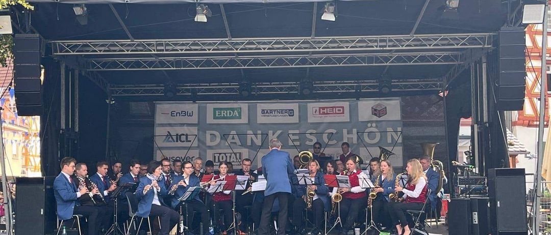 Ein Orchester spielt auf einer Freiluftbühne mit Vordach. Mehrere Sponsorenlogos sind auf dem Hintergrund zu sehen. Das Publikum sitzt vor der Bühne.