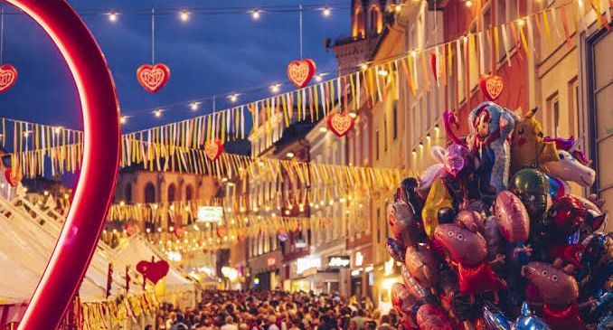 Eine überfüllte Straße mit bunten Luftballons und Herzdekorationen, unter einem Nachthimmel. Gebäude säumen die Straße, geschmückt mit Lichtern und Flaggen.